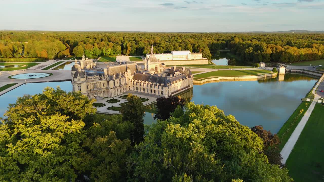 Aerial drone circling Château de Chantilly in France, surrounded by water and forest during sunset