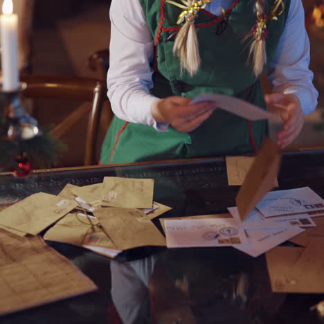 funny elf holding stack of envelopes for santa. dispersed young elf with letters. envelopes falling on the table from girl's hands on the christmas tree background.