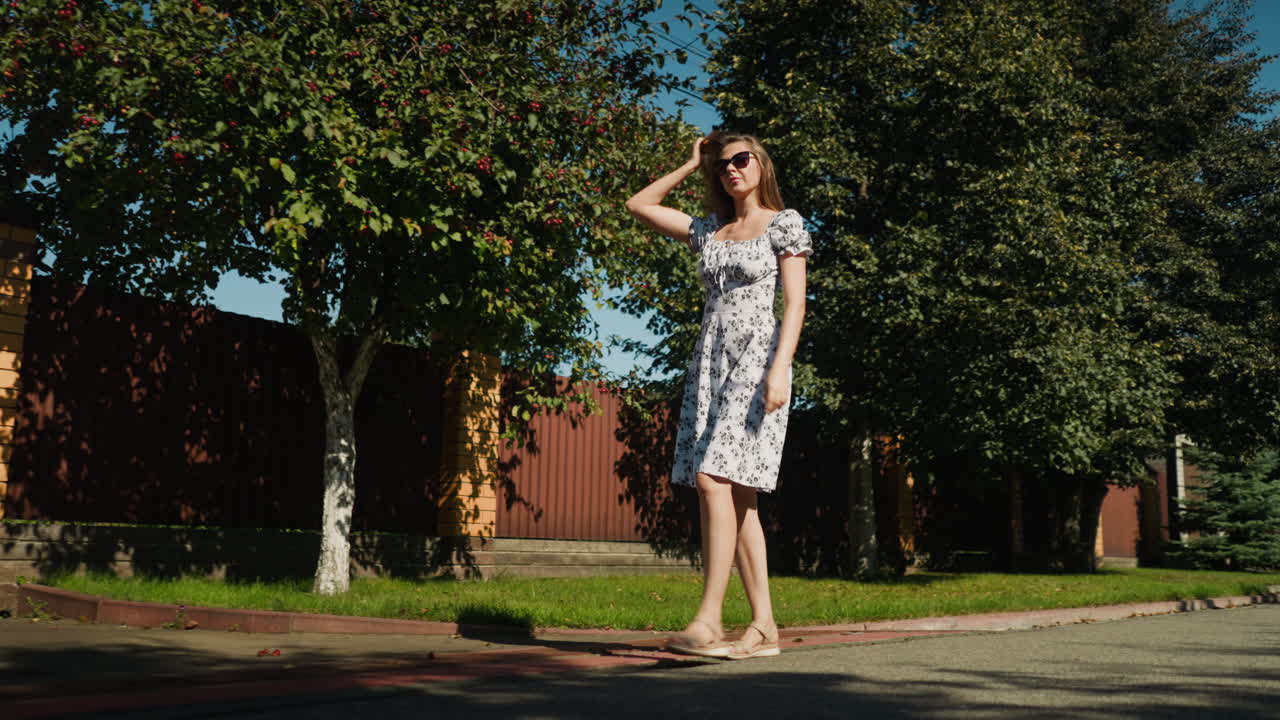 Confident young woman walking outdoors under bright sunlight and leafy tree shade, adjusting flowing hair to keep it off her glasses while flaunting her summer dress with relaxed poise