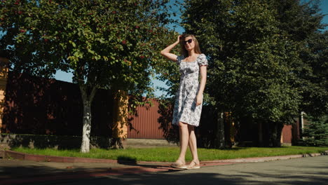 confident young woman walking outdoors under bright sunlight and leafy tree shade, adjusting flowing hair to keep it off her glasses while flaunting her summer dress with relaxed poise