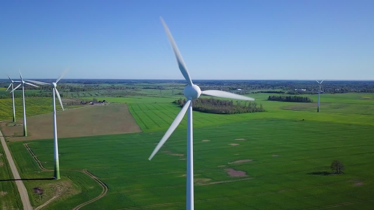 Aerial view of wind turbines generating renewable energy in the wind farm, sunny spring day, low flyover over green agricultural cereal fields, countryside roads, drone dolly shot moving left