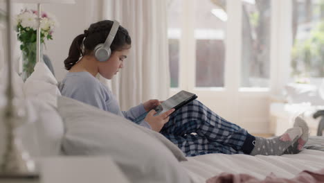 happy teenage girl listening to music wearing headphones browsing online using tablet computer relaxing on bed sharing lifestyle on social media enjoying weekend morning