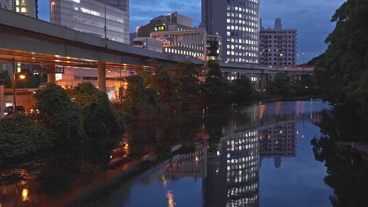The Benkei Bridge, a significant overpass, spans a tranquil canal at dusk, with modern buildings and reflections creating a dynamic urban night scene.