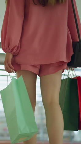 una dama con una camiseta rosa y pantalones cortos sostiene bolsas de compras en ambas manos mientras camina a través de un centro comercial brillantemente iluminado, los reflejos de luz rebotan en el vidrio, con otras personas que se ven moviendo