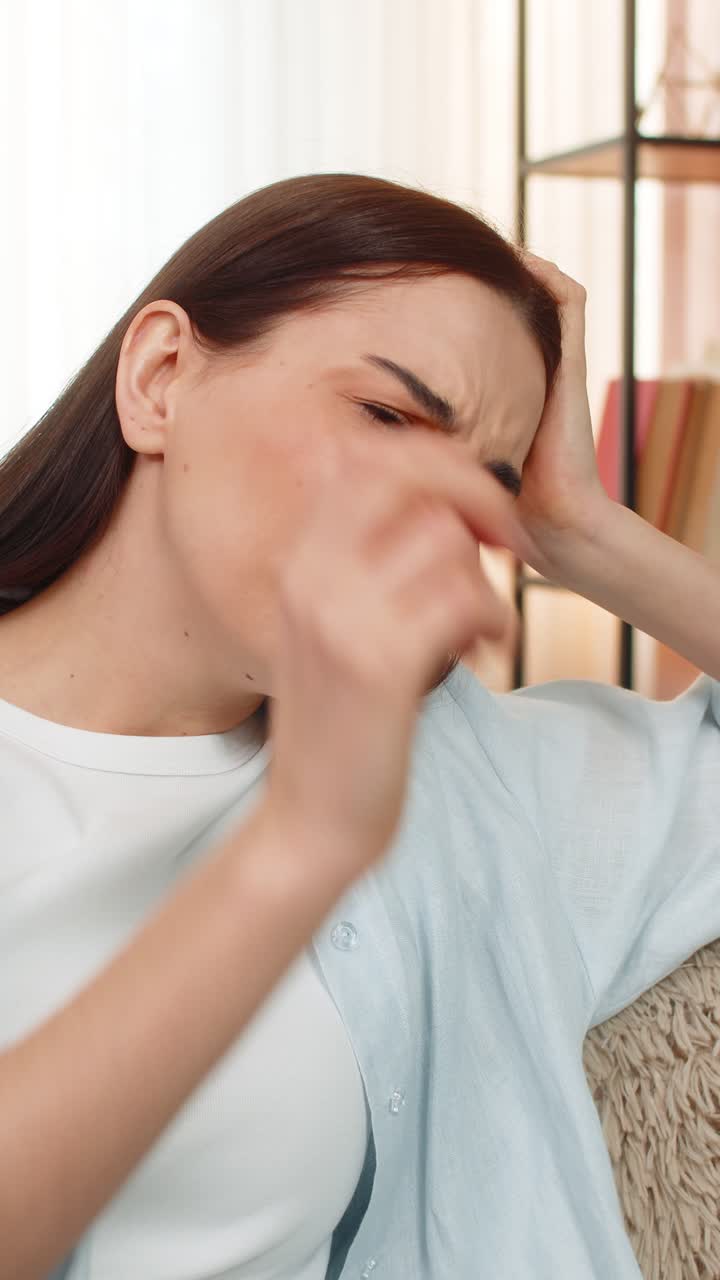 Young woman sitting on sofa holding head suffering from strong headache feeling uncomfortable pain