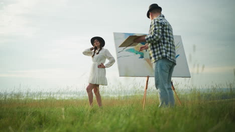 a sketch artist in a checkered shirt and jeans captures the image of a woman posing in a white dress and hat amidst a lush grassy field. the serene outdoor environment highlights the creative process