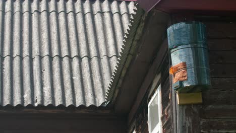 a cylindrical object resembling a beehive or insect trap attached to the wooden wall of a house with a corrugated roof.