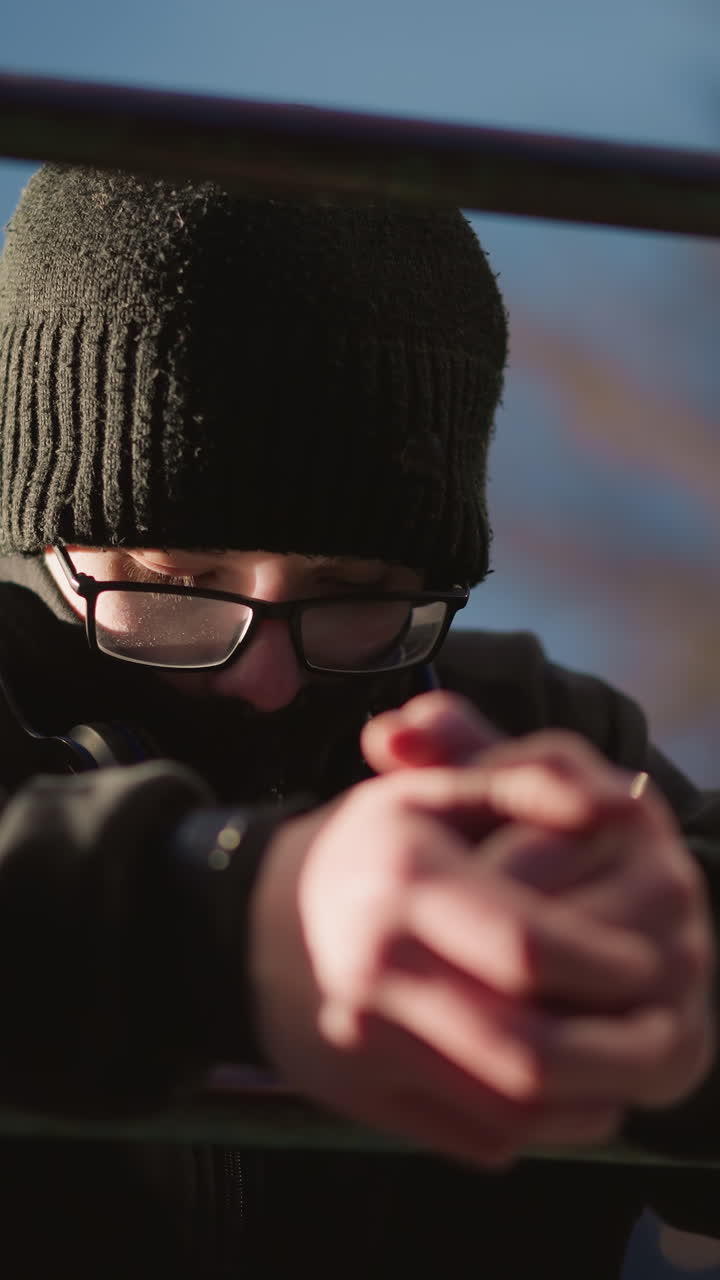 primer plano de un niño con gafas y una chaqueta negra, descansando en una barra de hierro con la cabeza inclinada, el fondo presenta luces borrosas y árboles desnudos