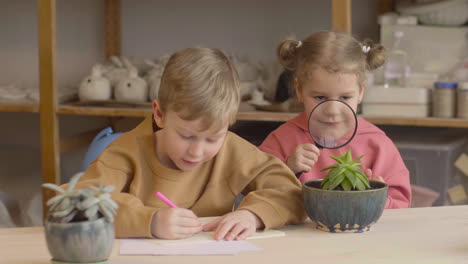 little girl observing a plant with a magnifying glass sitting at table near a friend who is drawing in a craft workshop