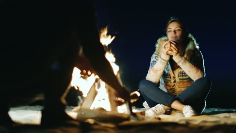 Friends-Relaxing-Around-The-Campfire-Woman-Drinking-Tea-From-A-Cup-A-Man-Chopping-Wood-With-An-Ax