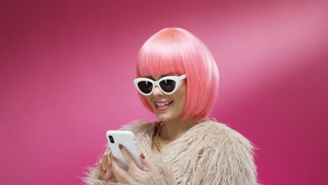 close up of cheerful young stylish woman in pink wig tapping and texting on mobile phone on colorful background and reading message