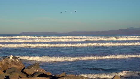 Capas-De-Olas-Rompiendo-Y-Rodando-Hacia-La-Costa-A-Lo-Largo-De-La-Costa-Durante-La-Hermosa-Puesta-De-Sol-Dorada-Con-Gaviotas-Sentadas-En-Primer-Plano,-Montañas-En-El-Fondo-Y-Pájaros-Volando-En-Un-Cielo-Azul-Claro-Y-Soleado