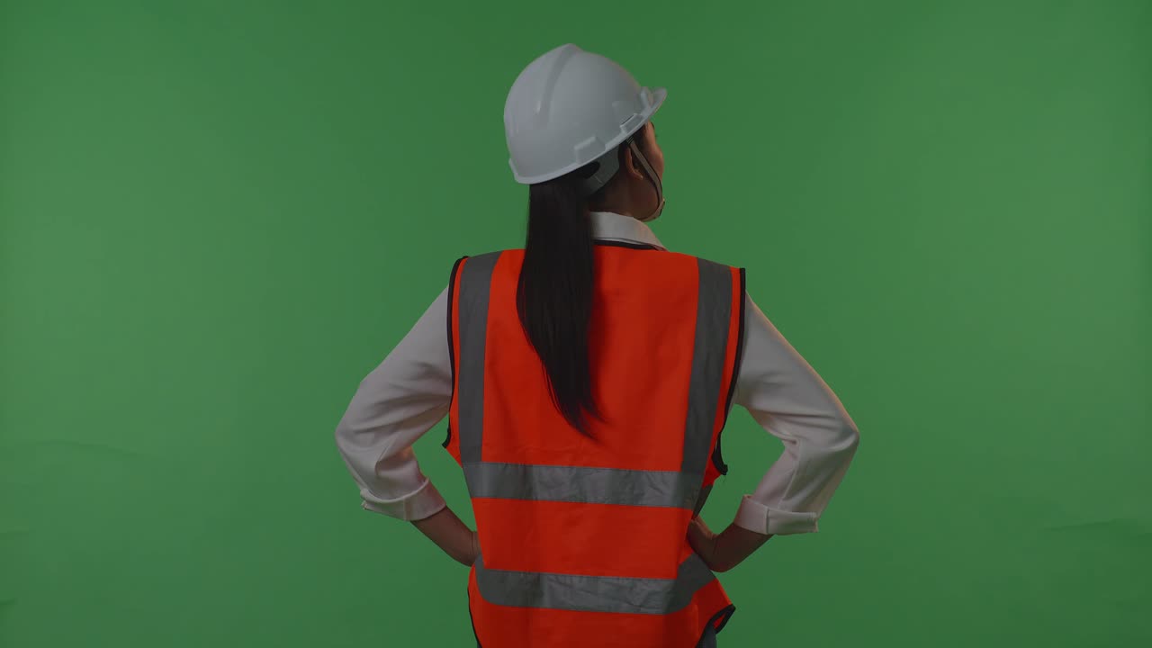 안전 헬을 착용한 여성 엔지니어가 녹색 스크린 배경 스튜디오에서 팔을 들고 서있는 동안 주위를 둘러보는 뒷면