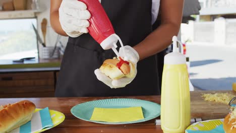 sección media de una mujer afroamericana con un delantal poniendo ketchup sobre un perrito caliente en un camión de comida
