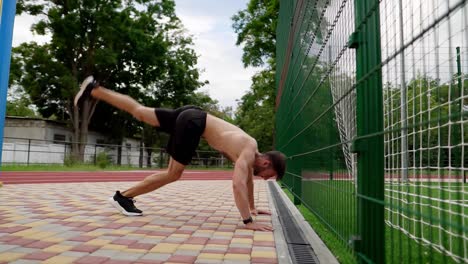 and-athlete-is-doing-push-ups-on-hands-standing-upside-down-near-the-wall-outdoors.-Young-man-topless-in-black-shorts