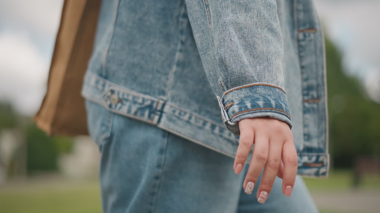 Primo piano di una giacca di jeans mentre si cammina in un parco, con luce naturale soffusa. Mano rilassata al fianco, texture di jeans blu vintage, passo lento e costante. Umore contemplativo, scatto lifestyle di un pomeriggio estivo che si concentra sul tessuto.