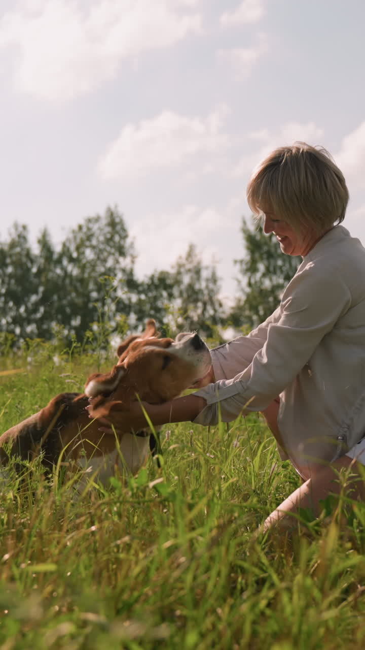 야외에 앉아있는 여자가 개 목을 문지르면서 개는 잔디에서 행복하게 귀를 흔들며 자연과 나무로 둘러싸여 있으며, 반려동물과 주인 사이의 장난스럽고 애정있는 유대를 포착합니다.