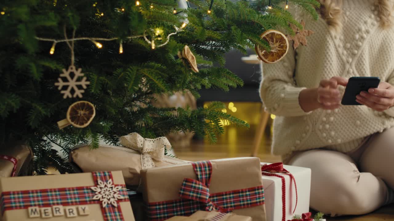 mujer caucásica irreconocible usando el teléfono sentada al lado del árbol de navidad.