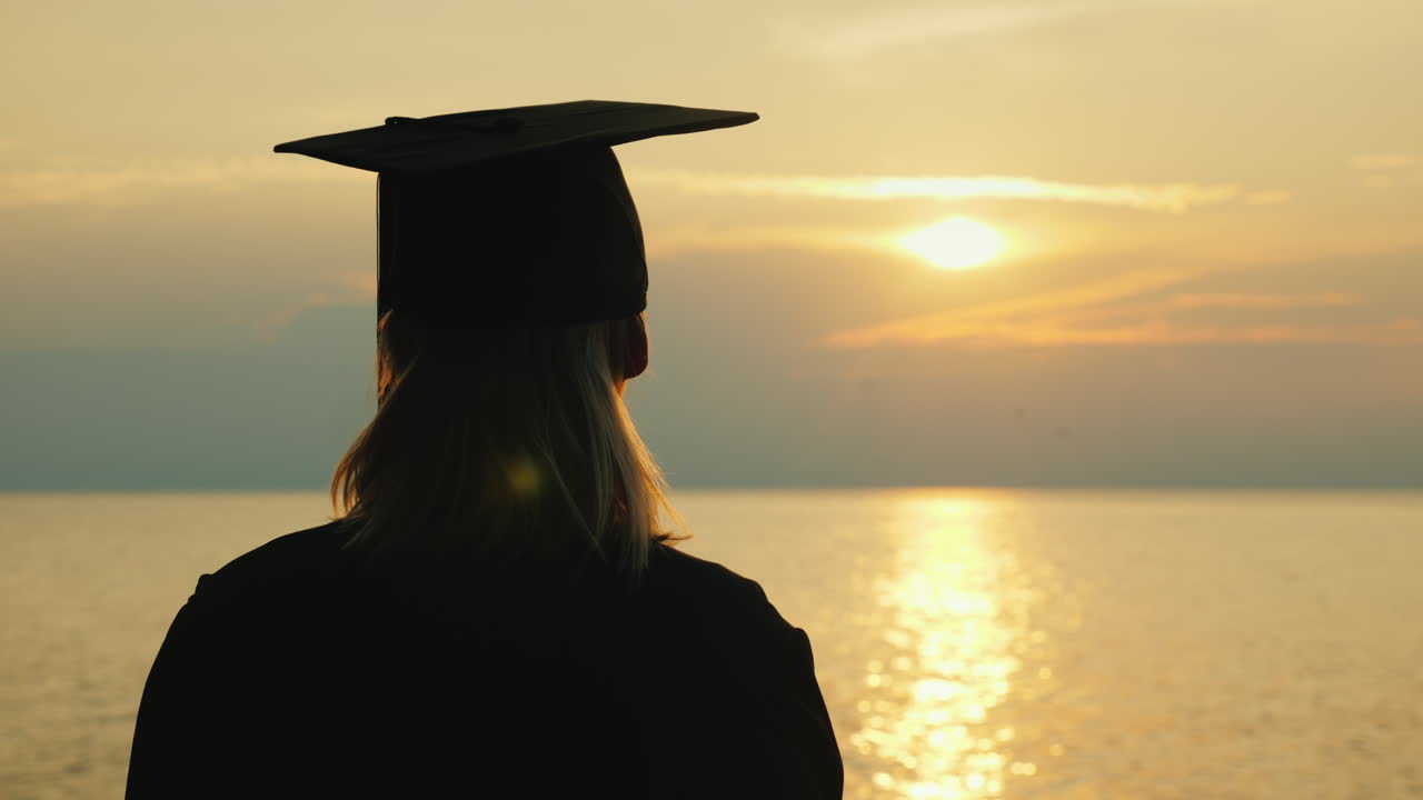 un soltero con un diploma en la mano y una gorra de graduado mira el amanecer sobre el mar