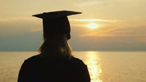 a bachelor with a diploma in hand and a cap of a graduate looks at the sunrise over the sea