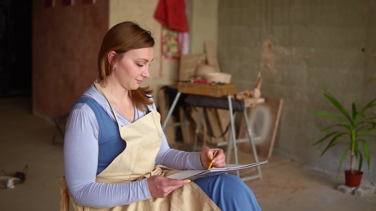 Artist woman at the workplace drawing pencil sketch. Long haired woman sitting on the chair at the workplace, holding work sheet on knees. Calm, inspirational work at art studio