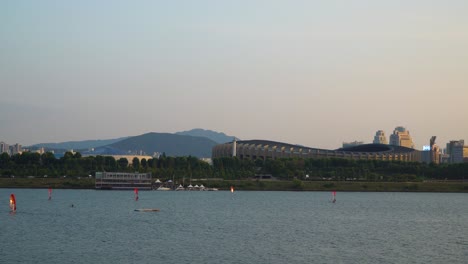 Gente-Haciendo-Windsurf-En-El-Río-Han-Al-Atardecer-Cerca-Del-Parque-Ttukseom,-El-Complejo-Deportivo-Jamsil-O-El-Estadio-Jamsil-Y-Montañas-Distantes-En-El-Fondo---Espacio-De-Copia-De-Gran-Angular