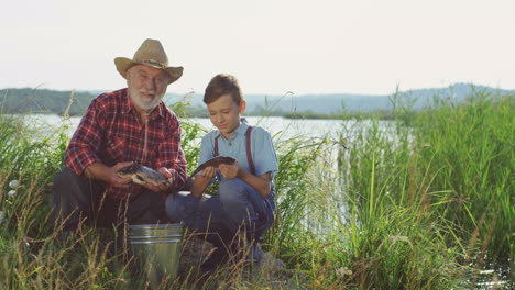 smiling old grandpa and his grandson looking at the camera holding fishes in the shore of a lake