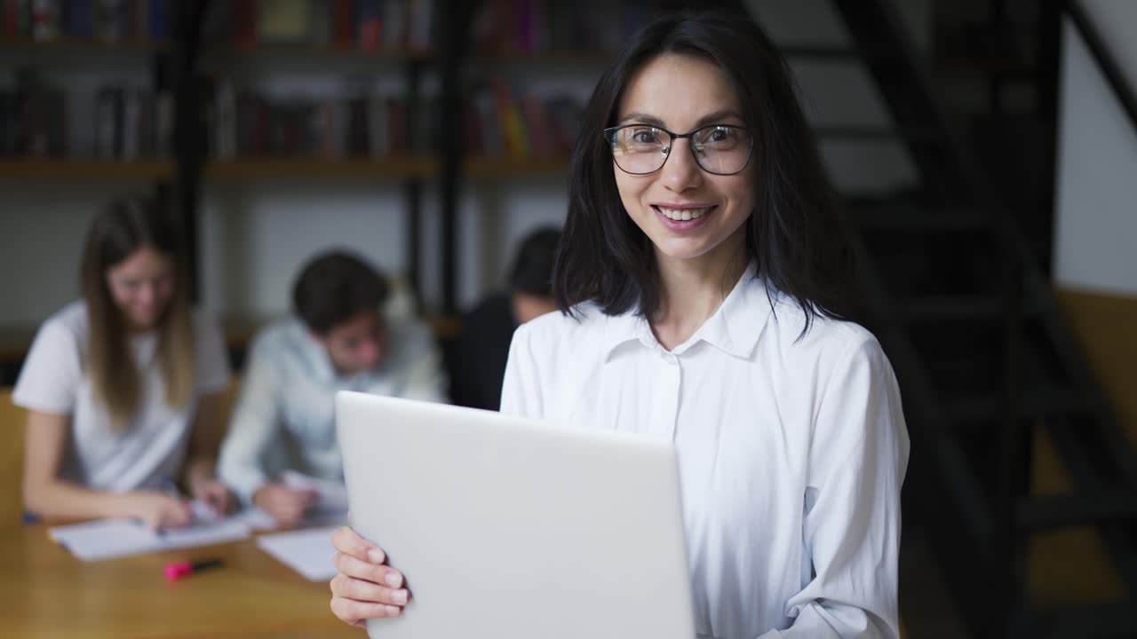 chica linda positiva con cabello negro en camisa, estudiante o maestro sosteniendo una computadora portátil y mirando a la cámara, sonriendo amistosamente