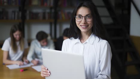 Positives,-Süßes-Mädchen-Mit-Schwarzen-Haaren-Im-Hemd,-Schülerin-Oder-Lehrerin,-Die-Einen-Laptop-In-Der-Hand-Hält-Und-In-Die-Kamera-Blickt-Und-Freundlich-Lächelt