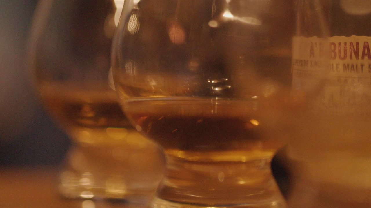 Multiple whiskey glasses with amber liquid are arranged in a row on a wooden bar, captured in soft, warm lighting with shallow depth of field