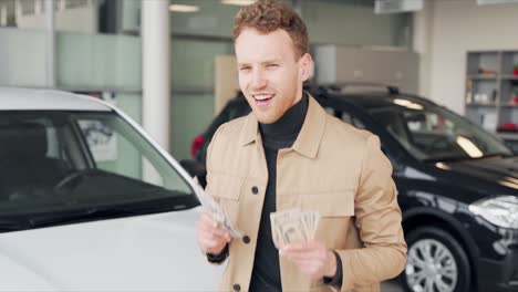 happy young man dancing with money in hands on the background of new cars