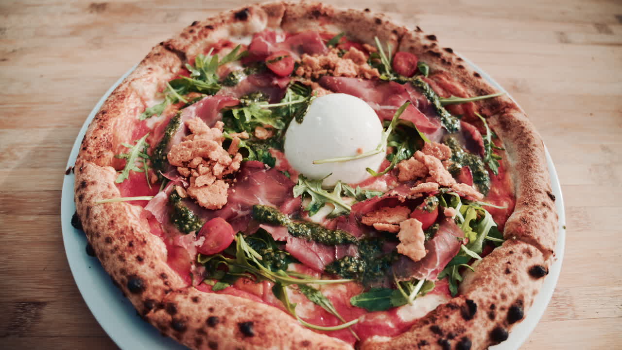 A beautifully shot overhead view of a gourmet pizza topped with burrata, prosciutto, cherry tomatoes, pesto, arugula, and crunchy crumbs