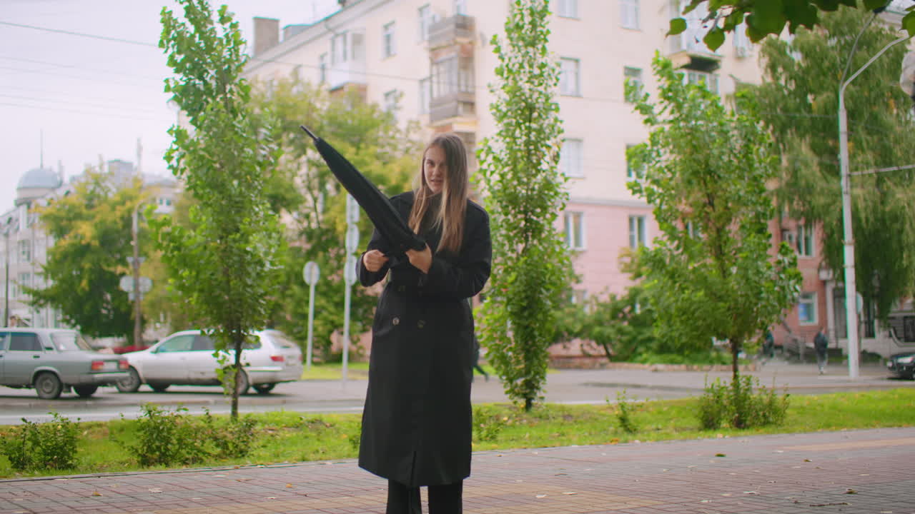 Woman in long black trench coat standing on city sidewalk holding closed umbrella, adjusting it with both hands while cars pass in background, surrounded by trees,apartment buildings in overcast weather