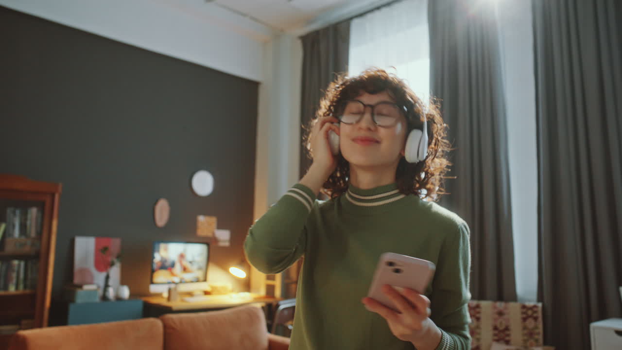 Carefree Asian Girl in Headphones Holding Phone and Dancing in Living Room