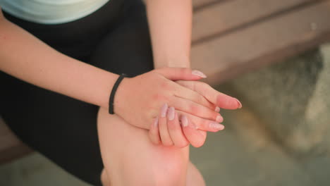 leg view of lady seated on wooden bench outdoors with leg crossed, gently clasping and rubbing hands together resting on knee, under warm sunlight, with background softly blurred