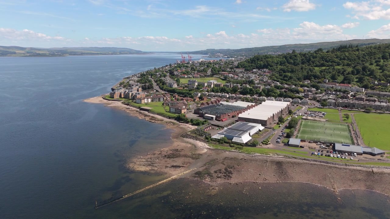 Un'ampia ripresa aerea cinematografica che mostra la suggestiva costa di Greenock, Scozia, con Battery Park, campi sportivi locali e il Firth of Clyde sotto un cielo azzurro brillante