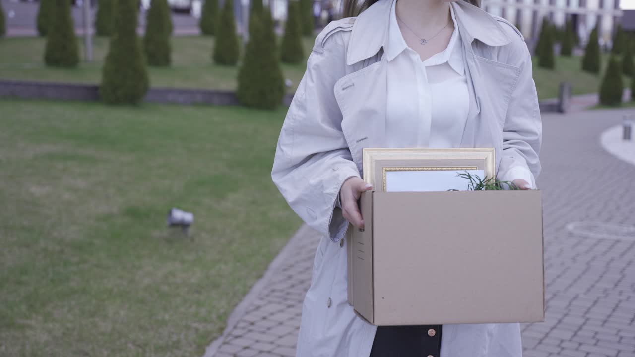사무실 건물 앞을 걷고 있는 예쁜 여자의 클로즈업. 그녀는 물건을 상자에 담아 가지고 다닙니다. 그녀는 해고되었습니다. 그녀는 슬프다.