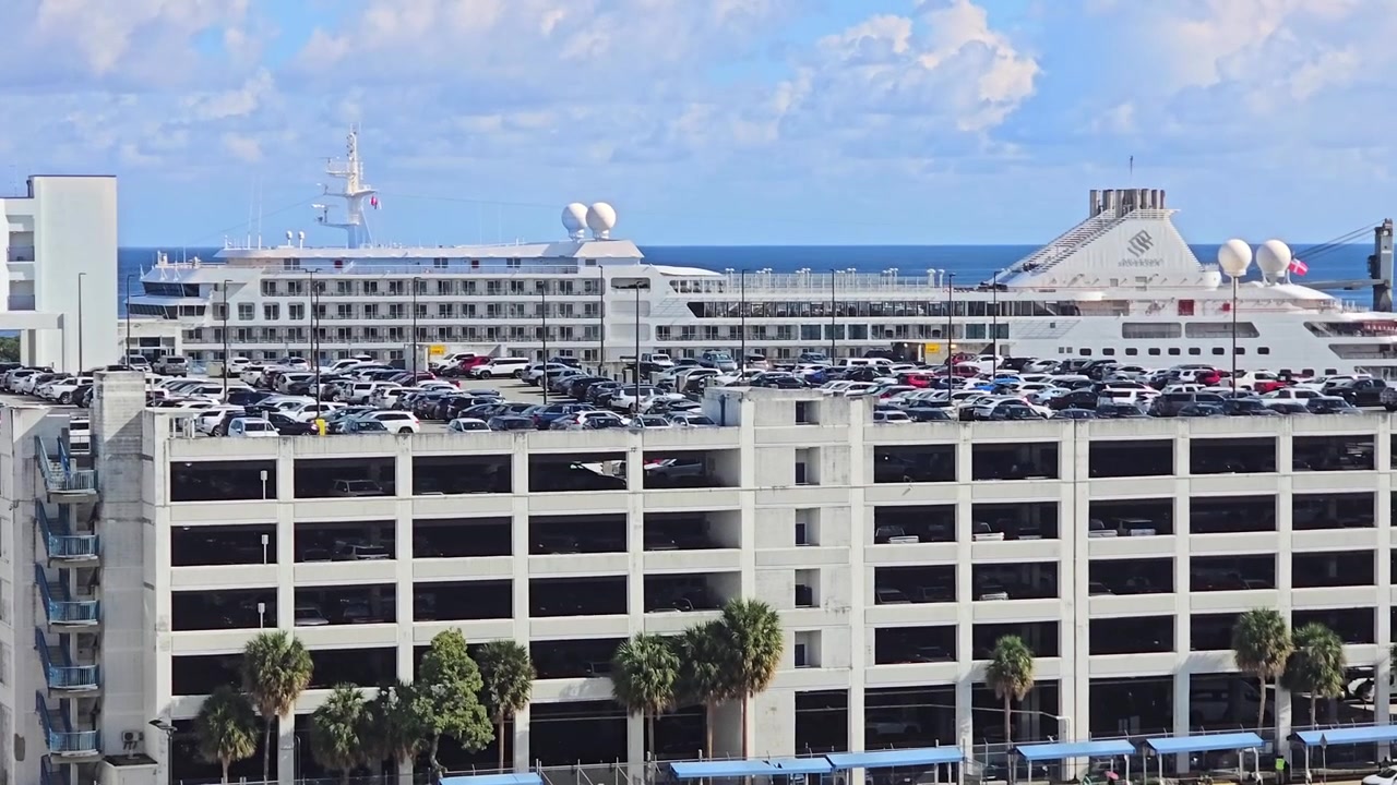 Una grande nave da crociera bianca è ormeggiata a un terminal portuale, direttamente dietro un enorme parcheggio multipiano affollato di auto, sotto un cielo azzurro