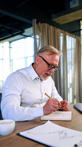 busy caucasian man working in the office. man sitting at desk uses laptop and taking notes. vertical video.