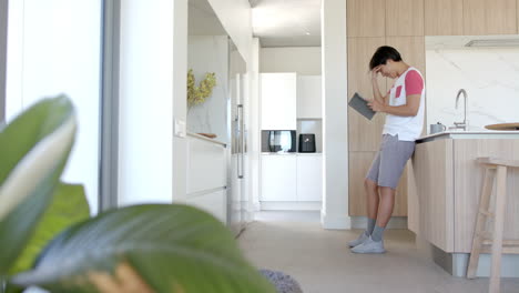 An-Asian-college-student-standing-in-kitchen,-looking-at-a-tablet