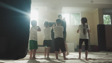 trainer demonstrates punching to kids at martial arts school. junior athletes repeat hits with hands following master in gym slow motion. healthy activity