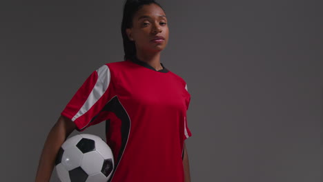 Studio-Portrait-Of-Serious-Female-Football-Or-Soccer-Player-Wearing-Team-Shirt-Holding-Ball-Under-Her-Arm-Shot-Against-Grey-Background