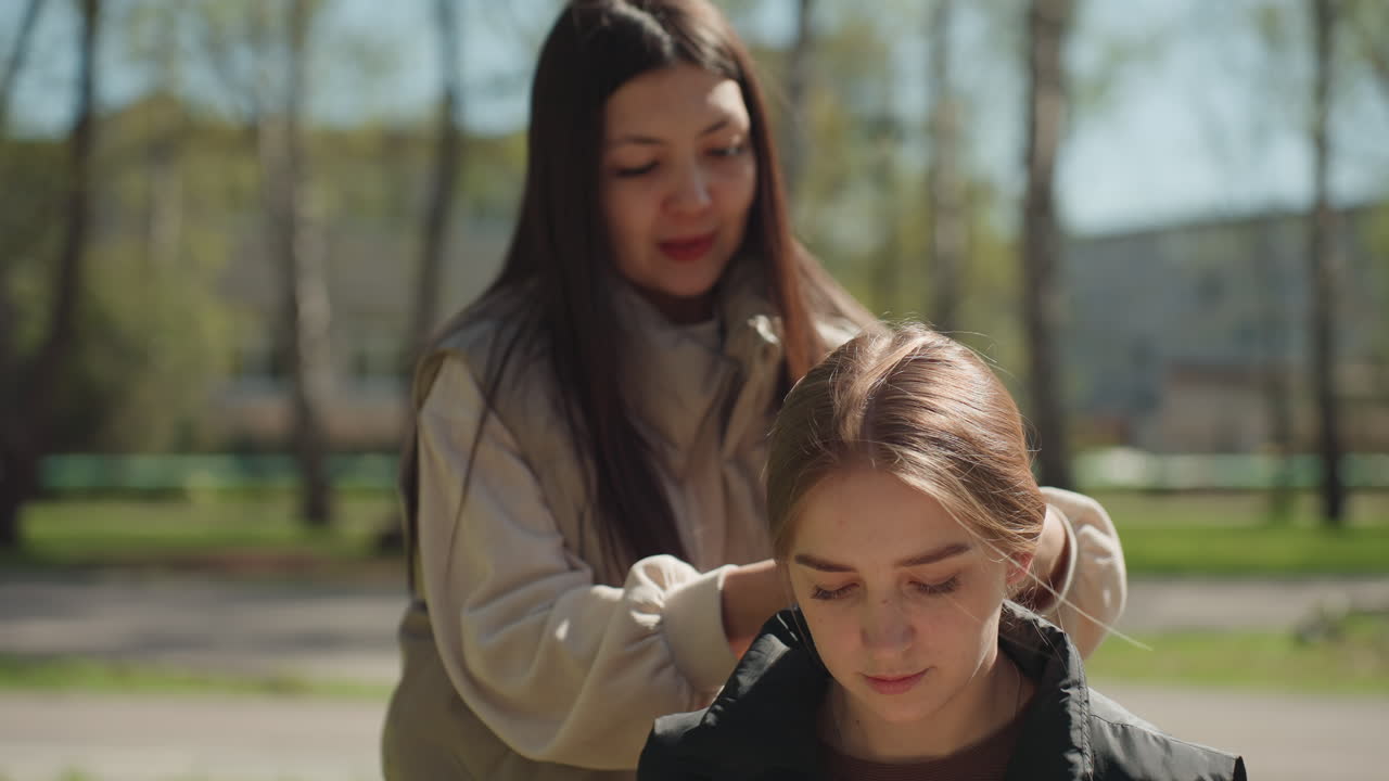 아시아 스타일리스트가 백인 친구의 머리를 야외에서 땋아주고 있어요. 손가락으로 긴 머리를 엮으면서 튜토리얼 같은 분위기를 연출하며 부드럽게 관리하고, 자작나무 사이로 햇빛이 비치는 가운데 캐주얼한 길거리 스타일링 및 그루밍 기술을 선보입니다.