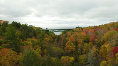 Drone-flies-over-green-bridge-towards-water-with-colorful-leaves-on-trees