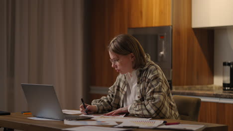 mujer con gafas trabajando de forma remota en una oficina en casa en un escritorio con una computadora portátil y notas de datos en un gráfico analista de economía de negocios