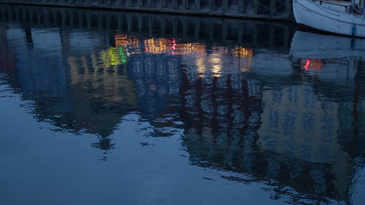 A close-up, artistic shot of the calm water in Copenhagen's Nyhavn canal at twilight. The dark blue surface of the water serves as a canvas for the shimmering, distorted reflections