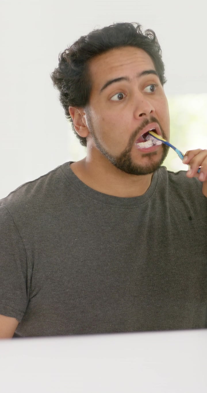 Vertical video: African American man brushing teeth at sink with blue brush foam, watching mirror