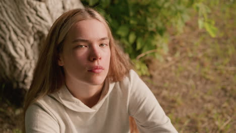 young lady seated on ground, looking up with focused expression, creating a contemplative mood, background features natural greenery and tree bark textures