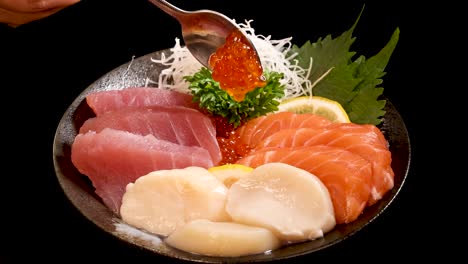 a hand uses a spoon to place salmon roe onto a bowl of assorted sashimi, including tuna, salmon, and scallop, under studio lighting with a black background