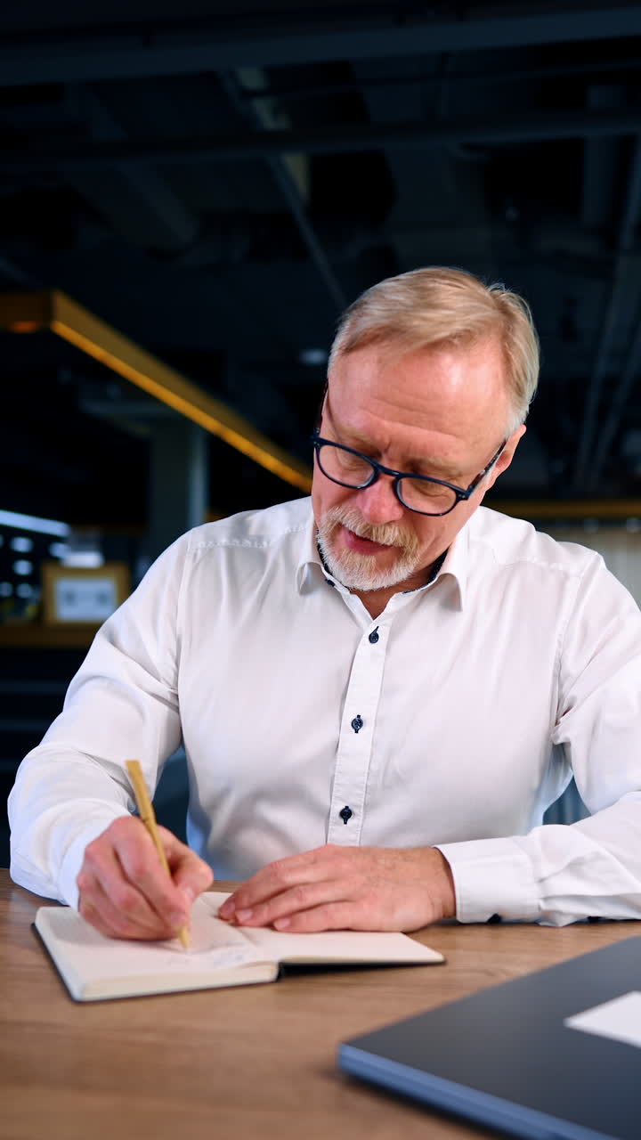 Puzzled man looks at his notebook on the desk. A sudden idea comes to man's mind and he writes it down. A writer, a businessman at work. Vertical video.